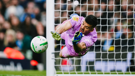 A goalkeeper in a purple jersey is diving to make a save. The soccer ball is flying towards the goal. The scene captures action during a match.