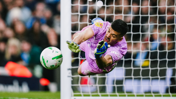 A goalkeeper in a purple jersey is diving to make a save. The soccer ball is flying towards the goal. The scene captures action during a match.