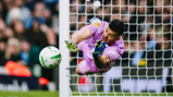 A goalkeeper in a purple jersey is diving to make a save. The soccer ball is flying towards the goal. The scene captures action during a match.