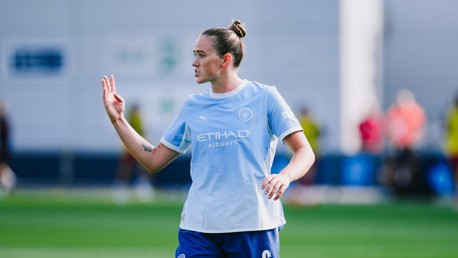 A football player wearing a Manchester City training kit gestures with their hand while on the field.