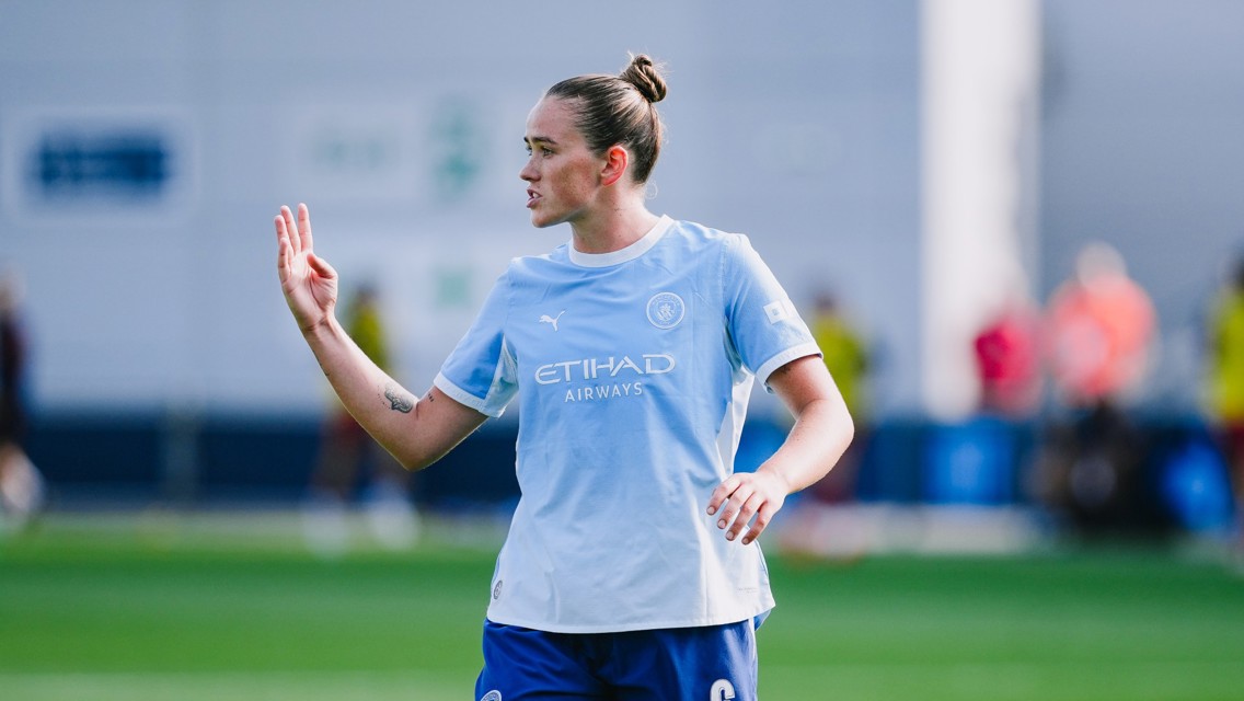 A football player wearing a Manchester City training kit gestures with their hand while on the field.