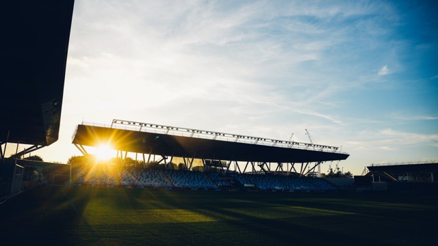 A stadium with empty seats and the sun setting behind a grandstand, casting shadows on the field.