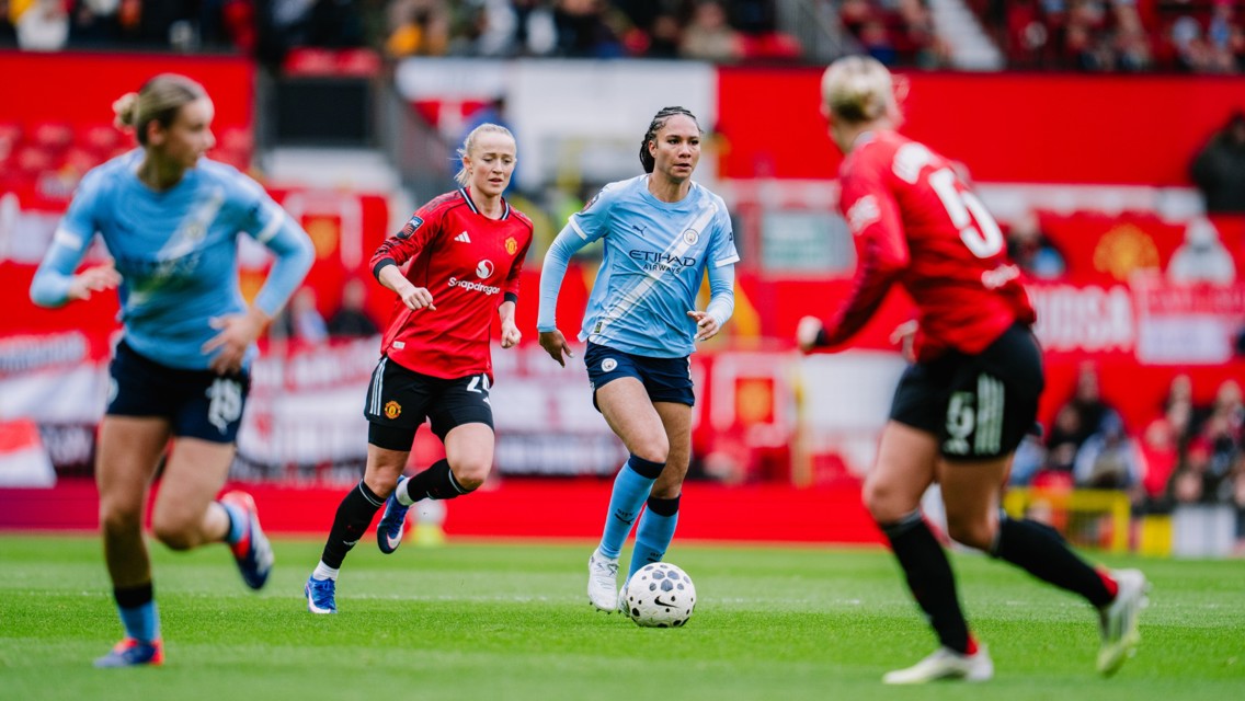Soccer players from two teams, one in red and the other in blue, in action during a match. The red team has a prominent logo and is facing off against the blue team on a grassy field.