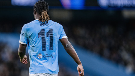 A football player wearing a Manchester City jersey with the name 'Doku' and number 11 on the back. The jersey shows the Manchester City logo and a community patch.