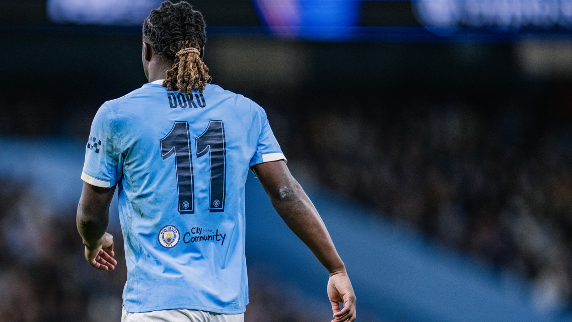 A football player wearing a Manchester City jersey with the name 'Doku' and number 11 on the back. The jersey shows the Manchester City logo and a community patch.